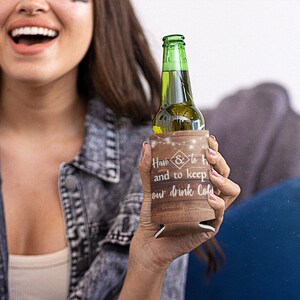 Op de afbeelding: Een bruine, houtlook-blikjeskoeler met witte tekst die "Have & to hold and to keep our drink cold" luidt. De blikjeskoeler heeft een string witte lampjes rond de bovenkant.