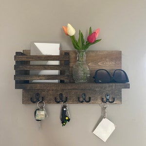 May include: A dark brown wooden key holder with four hooks, a small shelf, and a mail slot. There are keys, sunglasses, and a vase of pink and white tulips on the shelf.