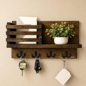 May include: A dark brown wooden wall-mounted organizer featuring a slatted mail slot, a flat shelf holding a small potted green plant, and four black metal double hooks. Three sets of keys are hanging from the hooks, and a white envelope is tucked into the mail slot.