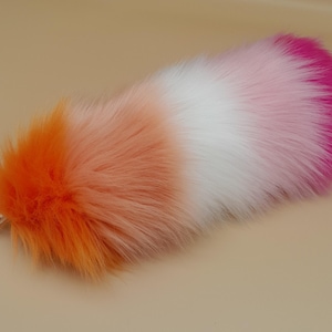 May include: A fluffy, faux fur tail in a gradient of colors: orange, peach, white, pink, and magenta. The tail has a silver-colored metal clasp attached to the base. The tail is designed to be worn as an accessory.