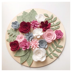 May include: A circular wooden base decorated with felt flowers and leaves. The flowers are in shades of pink, red, white, and grey. Green felt leaves are interspersed among the flowers, creating a floral arrangement.