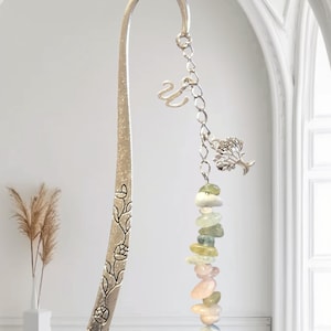 May include: Silver metal bookmark with floral detailing and a chain of colourful gemstones. The chain is adorned with a tree charm, a letter "W" charm, and a crescent moon charm. The bookmark is approximately 18 cm long.