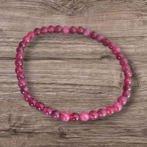 May include: A bracelet made of small, round, deep pink gemstones. The beads are strung together, forming a circular shape. The polished beads are set against a wooden background. The bracelet is approximately 18 cm in circumference.