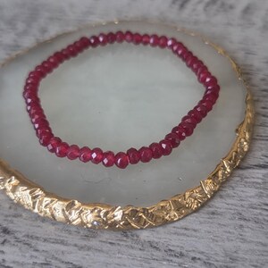 May include: A circular bracelet composed of small, faceted, deep red beads. The beads have a reflective, multi-faceted surface that catches the light, creating a consistent, uniform appearance throughout the entire length of the circular jewellery piece.