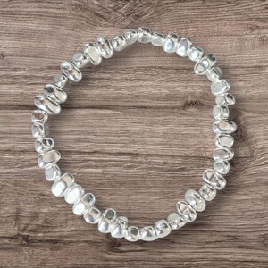 May include: A silver bracelet made of smooth, irregularly shaped beads. The beads are reflective, giving a polished look. The bracelet is circular and appears elastic, designed to stretch and fit the wrist. The bracelet is approximately 18 cm in circumference.