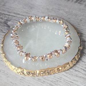 May include: A bracelet with silver and gold-coloured beads sits on a white and gold-edged decorative plate. The bracelet features small, irregular-shaped beads, giving it a textured look. The plate has a gold-coloured, textured edge.