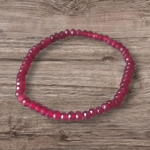 May include: A circular bracelet composed of numerous small, faceted, deep magenta-coloured beads. The beads are strung together in a continuous loop, displaying a uniform size and a reflective, multi-faceted surface texture throughout the entire piece.