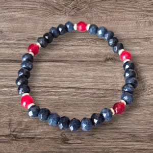 May include: A bracelet made of dark blue and red faceted beads. The beads are strung on a white cord, creating a repeating pattern. The bracelet is presented on a wooden surface.