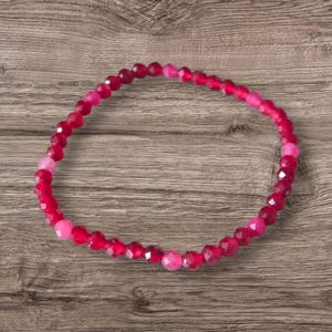 May include: A bracelet made of faceted, round beads in shades of red and pink. The beads are strung on an elastic band. The bracelet is displayed on a wooden surface. The beads have a polished, reflective surface.