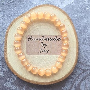 May include: A bracelet of peach-coloured, round beads. The bracelet is displayed on a small, round wooden slice. A small, rectangular tag with the words "Handmade by Jay" is placed on the wooden slice.