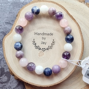 May include: A bracelet made with amethyst, rose quartz, white howlite, and blue sodalite beads. The bracelet is on a wooden surface with a tag that says "Handmade by Jay".