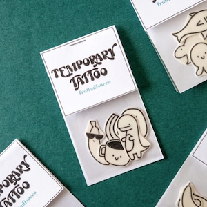 May include: Four white envelopes with black text that reads "Temporary Tattoo" and "fruttadisucco". Each envelope contains a different set of temporary tattoos. The tattoos are black and white and feature a banana, a coffee cup, a dinosaur, a fish, a turtle, and an apple.