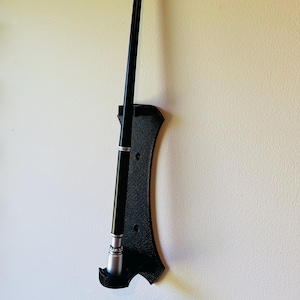 Pode incluir: Uma varinha preta com detalhes prateados, exibida em um suporte preto texturizado. A varinha é longa e fina, com um design elegante. O suporte está montado em uma parede clara, criando um contraste marcante.