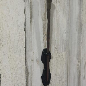 May include: A brown, slender, rod-shaped wand with a textured, bulbous handle, held vertically by a black, glossy, contoured wall-mounted display bracket. The wand features a tapered shaft and a detailed, organic-looking grip at the top end.