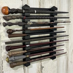 Könnte beinhalten: Eine Sammlung von elf aufwendig gestalteten Zauberstäben, die auf einem schwarzen, an der Wand befestigten Gestell ausgestellt sind. Die Zauberstäbe variieren in Farbe und Stil, mit dunkelbraunen, schwarzen und goldenen Akzenten. Das Gestell ist an einer hellen Holzpaneelwand befestigt.