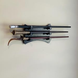 May include: Three decorative wands of varying designs are displayed on a black wall mount. The wands are black, brown, and silver, with intricate details. The wands are of different lengths and styles, showcasing a fantasy theme.