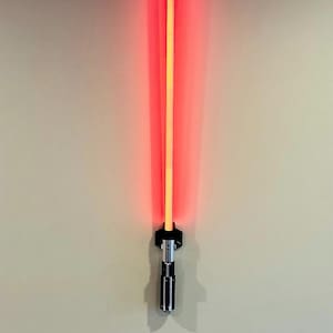 May include: A red and yellow lightsaber prop, mounted on a wall. The lightsaber features a black and silver handle with intricate details. The blade emits a bright red glow, creating a striking visual effect. The overall design is reminiscent of a science fiction weapon.