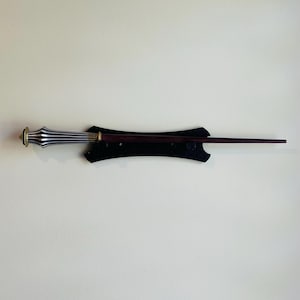 May include: A dark brown wooden wand with a silver and gold handle is mounted on a black wall mount.