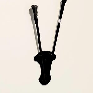 May include: Two black wands displayed on a black wall-mounted holder. The left wand features a textured, rough handle, while the right wand has a smooth, tapered shaft with a silver-colored ring detail near the top. Both are held in a vertical position.