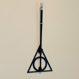 May include: A black wand with a silver tip and a Deathly Hallows symbol. The wand is hanging on a beige wall. The Deathly Hallows symbol is a triangle with a circle and a vertical line.