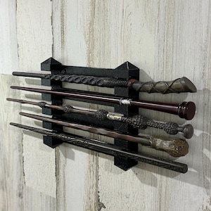 May include: A black wand display rack holding five detailed wands. The wands are various shades of brown and black, with intricate designs. The rack is mounted on a distressed, light-colored wooden wall.