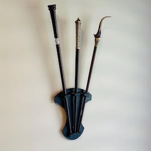Pode incluir: Um suporte de parede preto com três varinhas decorativas. As varinhas são feitas de madeira e metal com detalhes intrincados. O suporte tem um acabamento em madeira escura e um design simples.