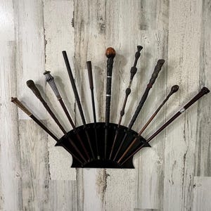 Puede incluir: Un soporte de pared negro en forma de abanico que sostiene diez varitas distintas de diversas longitudes, texturas y colores, incluyendo acabados en marrón oscuro, negro y metálico, dispuestas en un patrón semicircular.