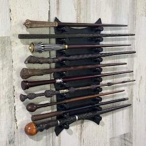 May include: A collection of ten distinct wands displayed horizontally on a black wall-mounted rack. The wands feature various designs, including metallic accents, carved wood textures, smooth finishes, and unique pommel shapes in shades of brown and black.
