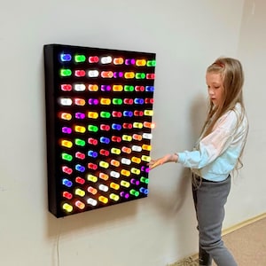 May include: A black rectangular LED light panel with many multi-coloured lights arranged in a grid. A young person stands beside the panel, their hand outstretched towards it. The panel is mounted on a light-coloured wall. The lights are in various colours.