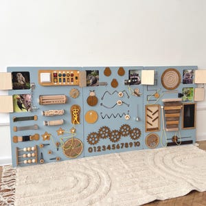 May include: A large, light blue sensory board with various wooden elements for tactile and visual exploration. Features include gears, abacus, animal images, and a number sequence from 0 to 10. The board is placed on a cream-colored rug.
