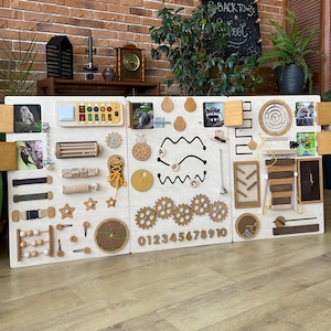 Peut inclure: Un tableau d'activités en bois avec différentes textures et activités pour les enfants. Il comprend une fermeture éclair, des boucles, un bouton, un système d'engrenages, un labyrinthe, une activité de laçage, un trieur de formes et une ligne de nombres. Le tableau est peint en blanc et a une finition en bois naturel.