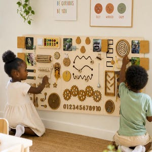 Op de afbeelding: Een groot, lichtgekleurd houten activiteitenbord voor aan de muur met diverse interactieve elementen zoals tandwielen, een doolhof, schakelaars, veters en de cijfers 0 tot en met 9. Het bord bevat kleine dierenfoto's en tactiele onderdelen.