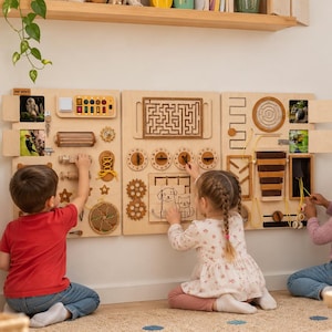 Op de afbeelding: Drie lichtgekleurde houten activiteitenborden aan een muur, voorzien van diverse interactieve elementen zoals doolhoven, tandwielen, klokken, grendels en dierillustraties. De borden zijn ontworpen voor tactiel spel en zintuiglijke verkenning.