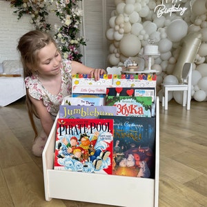 May include: White wooden bookshelf with a variety of children's books. The books have colorful covers with illustrations and titles such as "Little Grey Rabbit", "The Three", "Jumblebum", "збука", "Plunge into the Pirate Pool", and "Чудовищная Сказка".