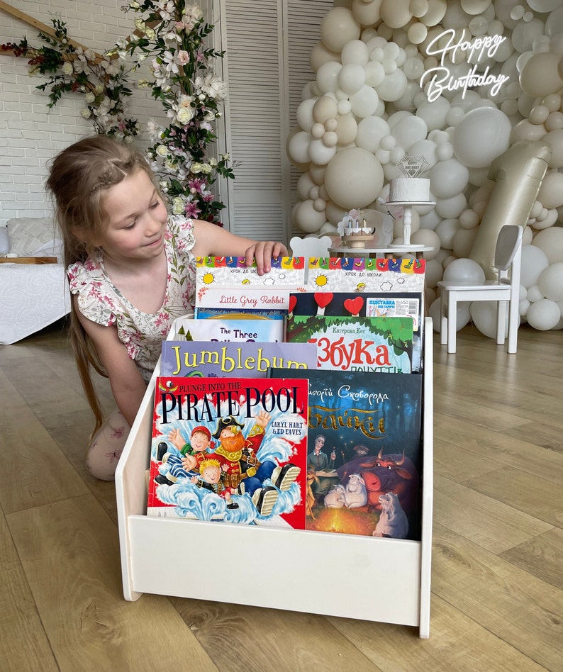 May include: White wooden bookshelf with a variety of children's books. The books have colorful covers with illustrations and titles such as "Little Grey Rabbit", "The Three", "Jumblebum", "збука", "Plunge into the Pirate Pool", and "Чудовищная Сказка".