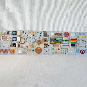 May include: A large, gray sensory board with various interactive elements for children. Features include gears, a rainbow, a clock, a maze, and images of planets and landscapes. Wooden blocks and colorful shapes are also incorporated.