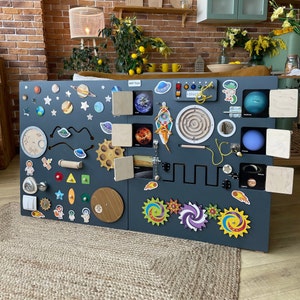 Op de afbeelding: Een houten activiteitenbord met een ruimtethema. Het bord is donkergrijs geverfd en heeft verschillende texturen, vormen en kleuren. Het bevat een grendel, een doolhof, een tandwiel, een schuifregelaar, een spiegel en een verscheidenheid aan andere interactieve elementen. Het bord is versierd met planeten, sterren en raketten.