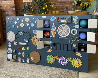 May include: A wooden activity board with a space theme. The board is painted dark gray and features various textures, shapes, and colors. It includes a latch, a maze, a gear, a slider, a mirror, and a variety of other interactive elements. The board is decorated with planets, stars, and rockets.