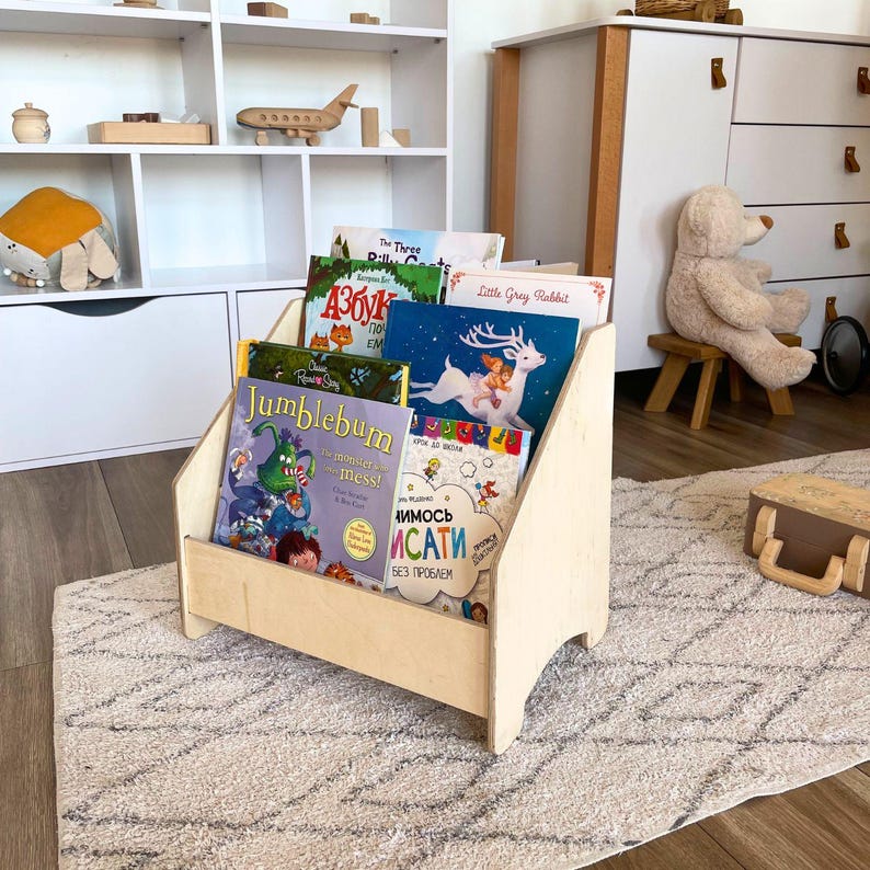 May include: A wooden bookshelf with five shelves, each holding a different children's book. The books are titled "The Three Billy Goats Gruff", "Азбука", "Little Grey Rabbit", "Jumblebum", and "ИМОСВ ИСАТИ БЕЗ ПРОБЛЕМ".