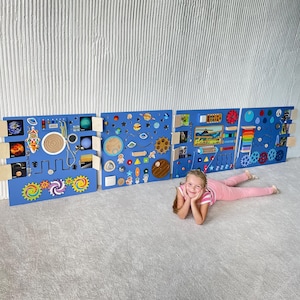 Op de afbeelding: Drie blauwe houten activiteitenborden met verschillende texturen, vormen en kleuren. De borden zijn aan een muur bevestigd en hebben een verscheidenheid aan interactieve elementen, waaronder tandwielen, knoppen en een doolhof. Een jong meisje ligt op de vloer voor de borden.