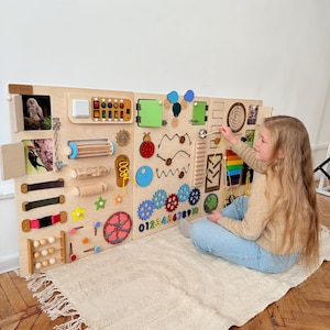 Op de afbeelding: Een groot houten activiteitenbord met diverse interactieve elementen voor kinderen. Bevat schakelaars, tandwielen, kralen en een kleine telraam. Het bord is lichtbruin met kleurrijke accenten en is ontworpen om de zintuigen en fijne motoriek te stimuleren.
