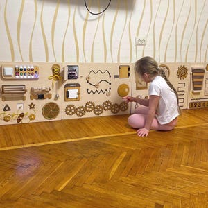 Op de afbeelding: Een houten activiteitenbord met verschillende texturen, vormen en kleuren. Het bord heeft een schuifdeur, een rits, een knop, een grendel, een tandwielsysteem, een doolhof en een getallenlijn. Een kind is aan het spelen met het bord.