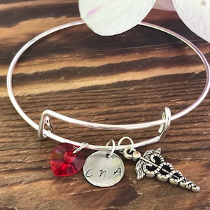 May include: Silver charm bracelet with a red heart charm, a round charm with the letters "CNA" and a silver charm of a caduceus.