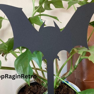 May include: Black metal bat silhouette garden stake. The stake is inserted into a potted plant with green leaves.