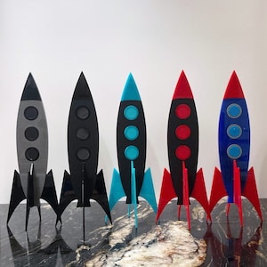 May include: Five rocket-shaped figurines, each with a different color scheme. The rockets are made of a black material and have three fins. The colors are gray, black, blue, turquoise, and red.