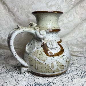 May include: A ceramic pitcher with a speckled cream and white glaze, featuring a dark brown rim and decorative brown accents. The vessel has a wide, flared neck, a rounded body, and a prominent, ornate handle with curled, sculptural details.