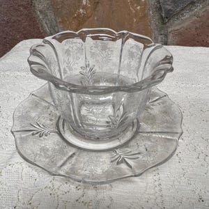 May include: A clear glass bowl with a scalloped rim sits atop a matching plate. Both pieces feature an etched floral design. The bowl has a flared shape, and the plate has a scalloped edge. The set is likely vintage.