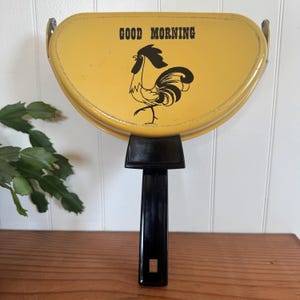 May include: Yellow vintage pancake griddle with a black handle. The griddle has the words "GOOD MORNING" and a rooster illustration in black. The griddle is on a wooden surface.