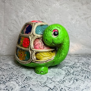 May include: A ceramic turtle figurine with a green head and legs, and a shell decorated with colorful floral patterns. The shell is divided into sections with a cream-colored border. The turtle has a smiling face and large, expressive eyes.