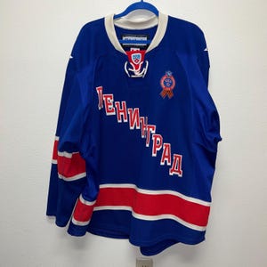 May include: A blue hockey jersey featuring the Cyrillic text LENINGRAD in red and white lettering across the chest. The jersey includes a white lace-up collar, red and white horizontal stripes on the sleeves and hem, and a circular commemorative patch.